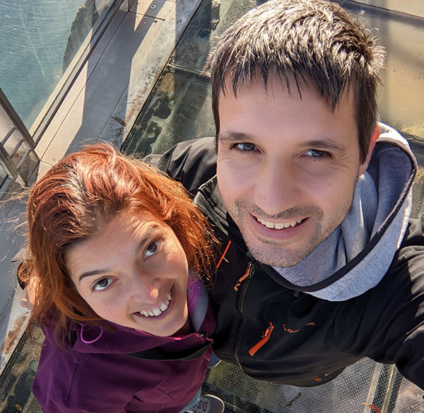 Um homem e uma mulher sorridentes estão tirando uma selfie em uma passarela com fundo de vidro. O homem tem cabelo curto e escuro e está vestindo uma jaqueta preta com zíper laranja. A mulher tem cabelo ruivo de comprimento médio e está vestindo uma jaqueta roxa. Eles estão olhando para a câmera.
